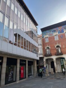 La imagen muestra un edificio moderno de cristal junto a una construcción tradicional con detalles arquitectónicos. Esta mezcla de estilos refleja la diversidad cultural y arquitectónica de la zona. La luz del día resalta los detalles de ambas edificaciones.